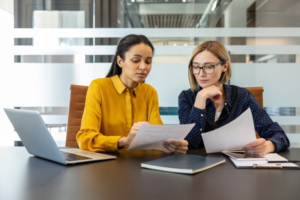 Two,Diverse,Businesswomen,Reviewing,Documents,And,Discussing,Work,,Collaborating,On